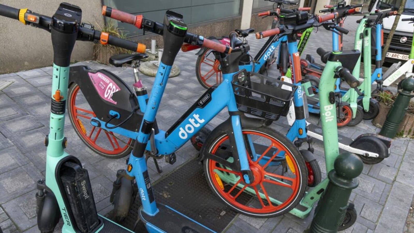 Shared electric scooters and a hire bicycle from several different operators parked together on a Brussels pavement.