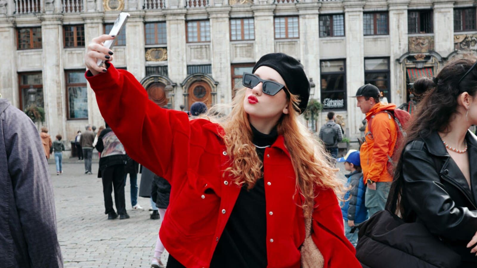 A visitor in a red jacket and beret takes a selfie in front of the guild houses on the Grand Place in Brussels, with other people crossing the square behind her.